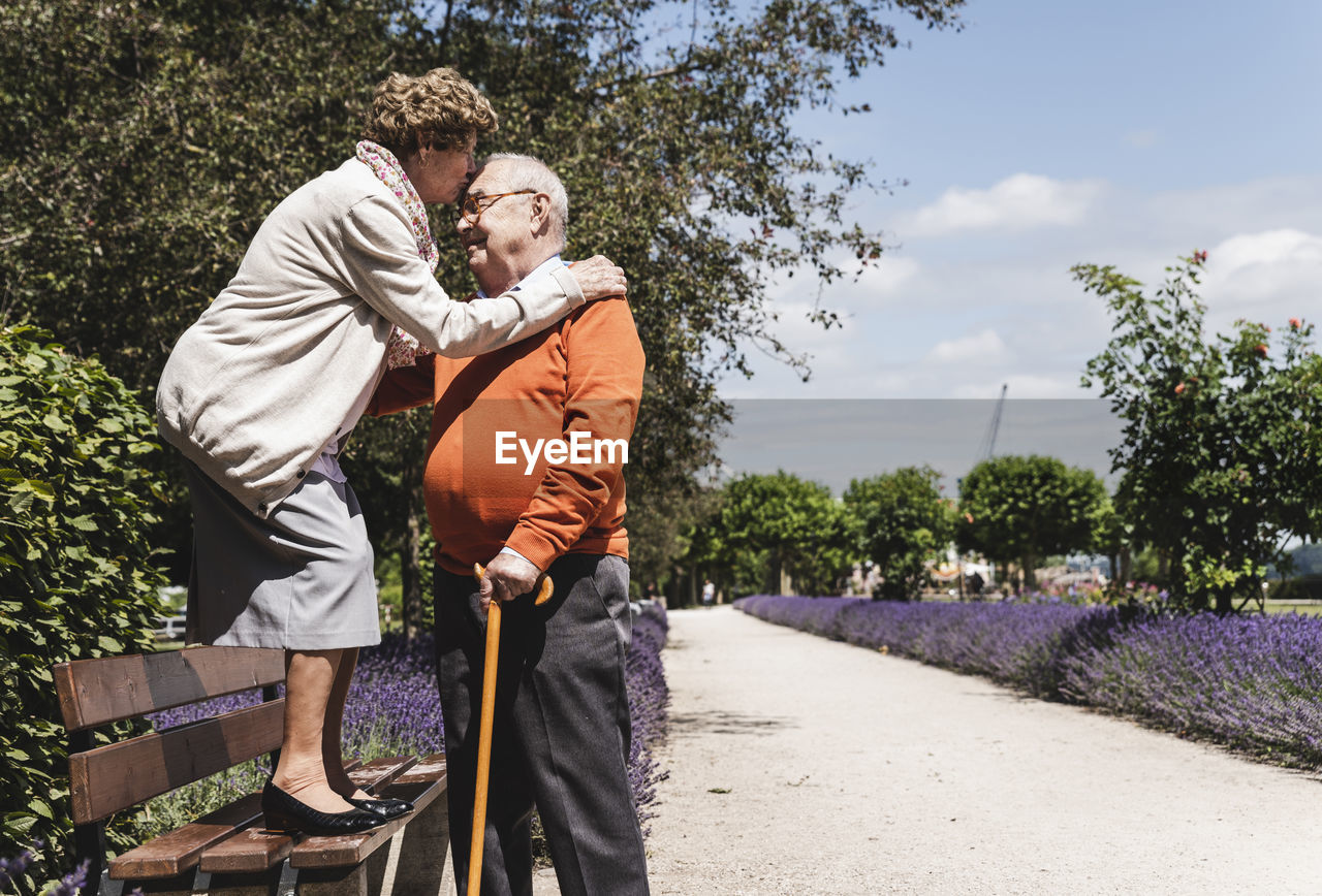 Senior couple having fun in the park, woman standing on bench kissing senior man on forehead