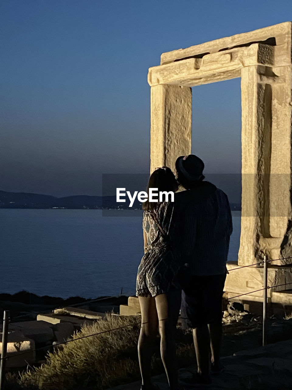 REAR VIEW OF COUPLE LOOKING AT SEA