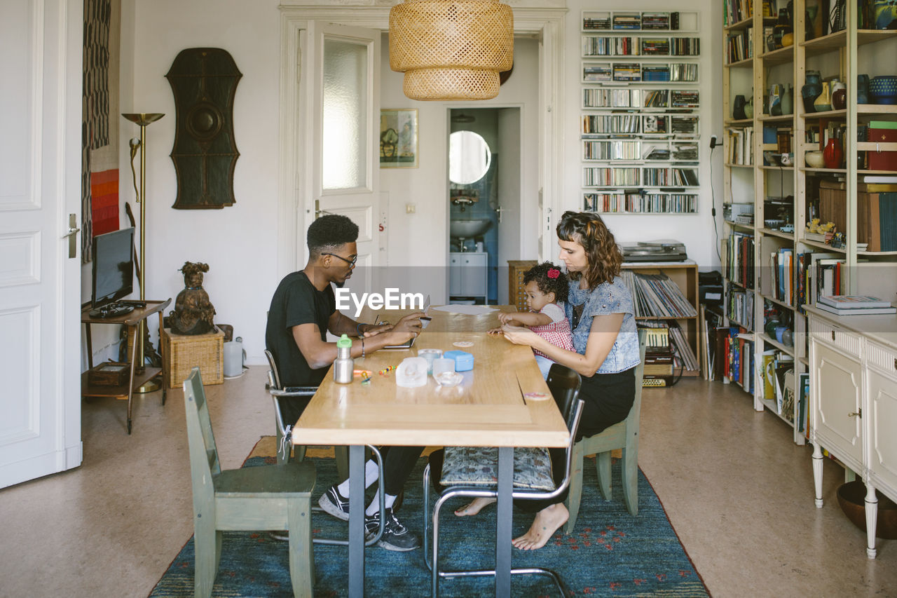 Young man using laptop while woman playing with daughter at dining table in house