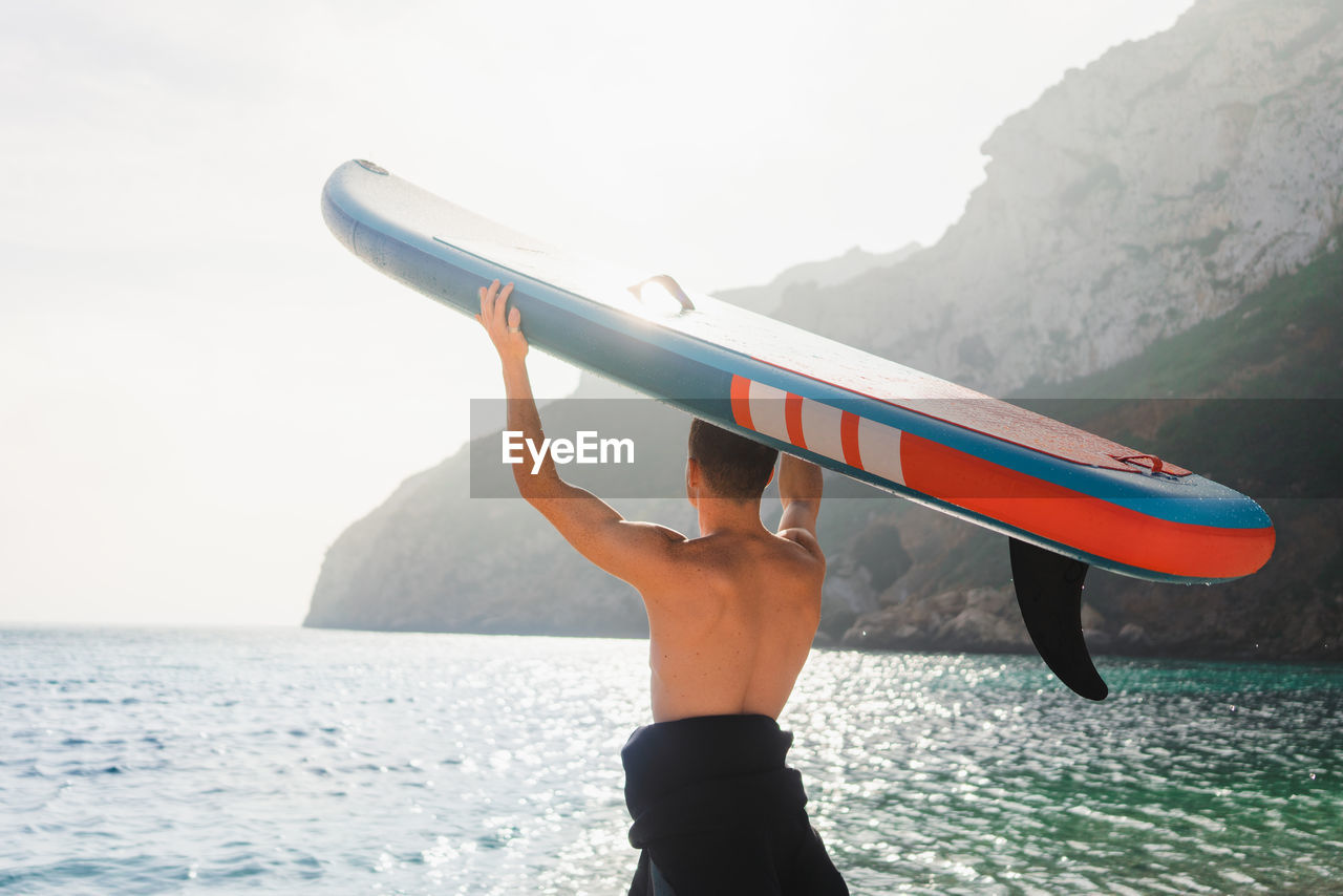 Back view of unrecognizable male surfer with sup board for paddle surfing standing on pebble coast of turquoise seawater in morning