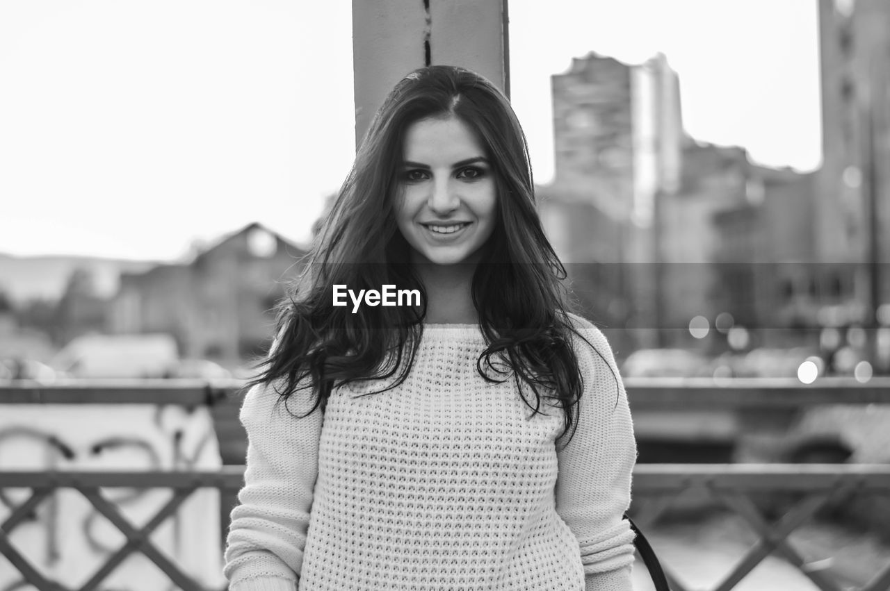 Portrait of smiling young woman standing against sky in city