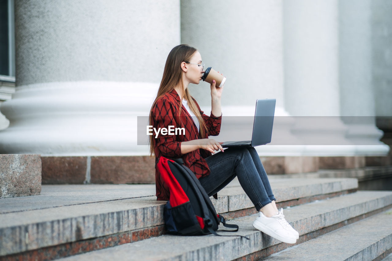 Outdoor portrait of female student with coffee cup and laptop. girl student has coffee break after