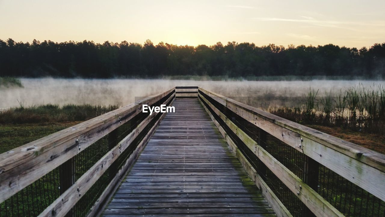 Wooden bridge over lake against sky during sunset