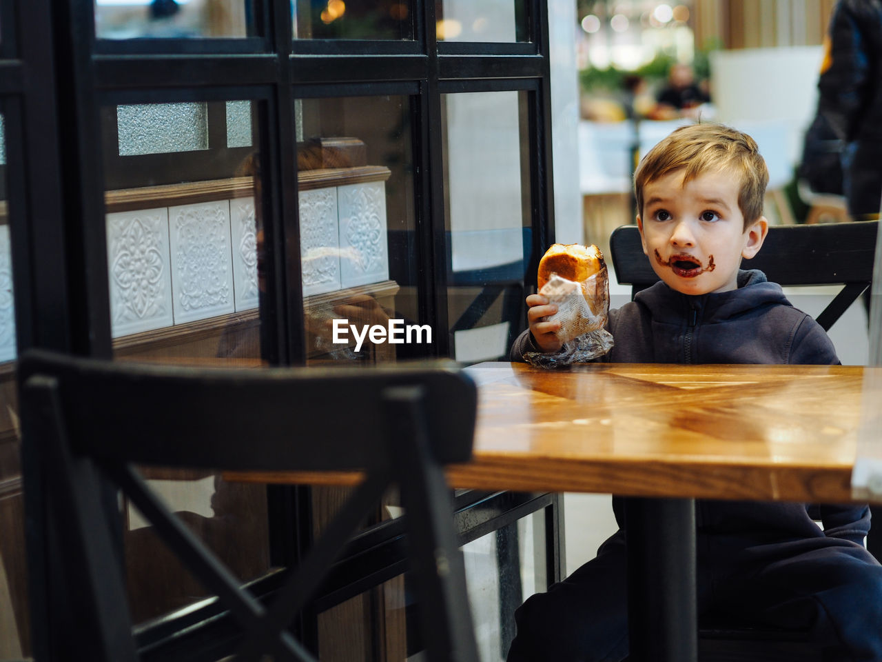 Toddler boy in casual clothes eating a chocolate bun in a food court there is a chocolate on face