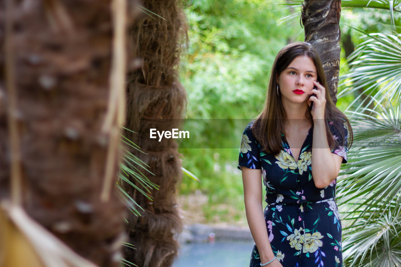 Beautiful young woman using phone while standing against tree trunk