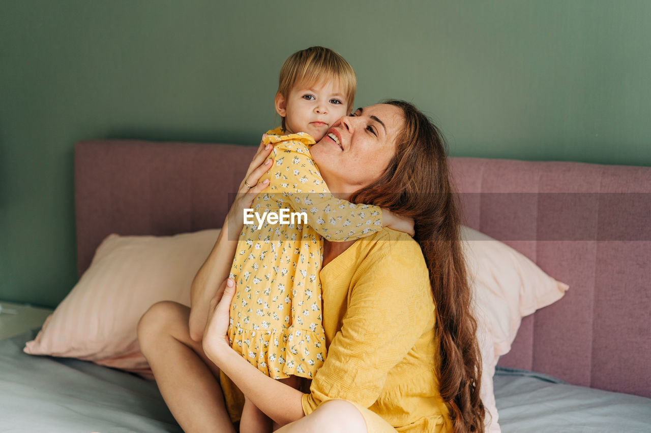 Red-haired mother hugs her little toddler daughter while sitting in bed. 