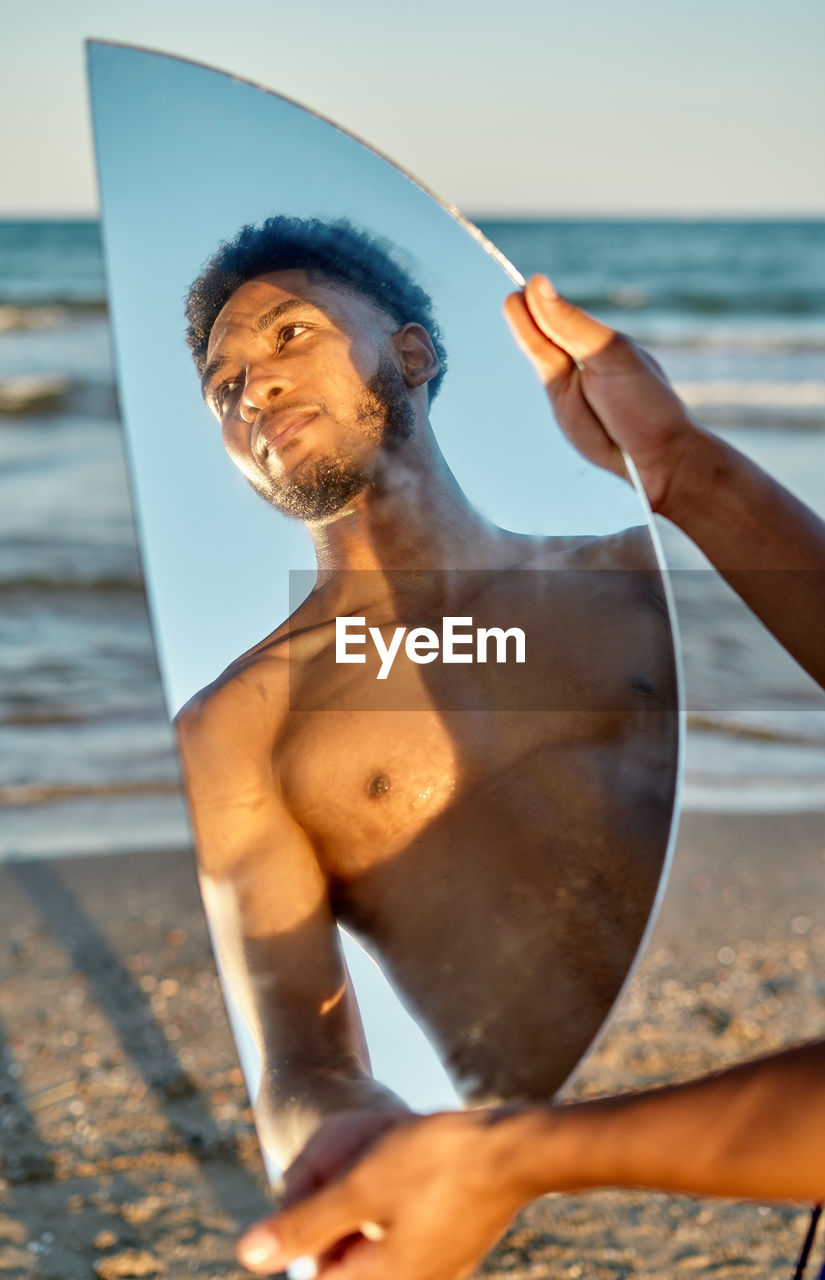 Crop african american male holding semicircular mirror while spending time on beach near sea