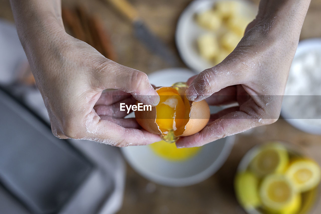 From above top view unrecognizable female breaking fresh chicken egg into bowl while cooking pastry in a wooden table with fresh ingredients