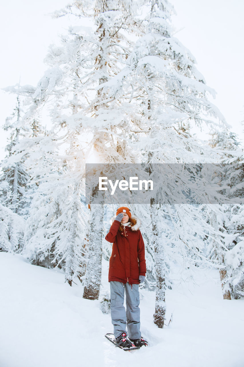 Full body woman in outerwear with snowshoes covering eye while standing near snowy spruces on cold winter day in valley of the ghosts in monts valin national park in quebec, canada