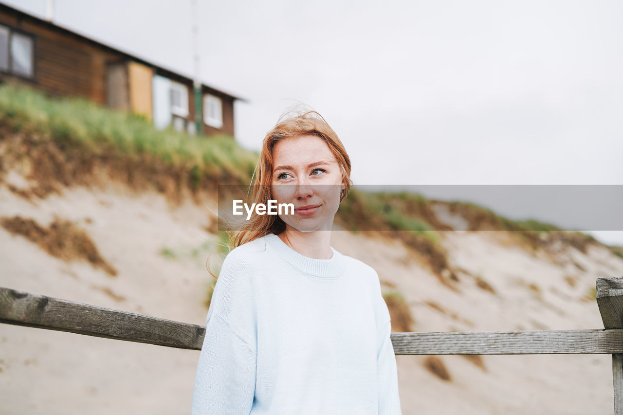 Portrait of young red haired woman in light blue sweater on sand beach by sea in storm