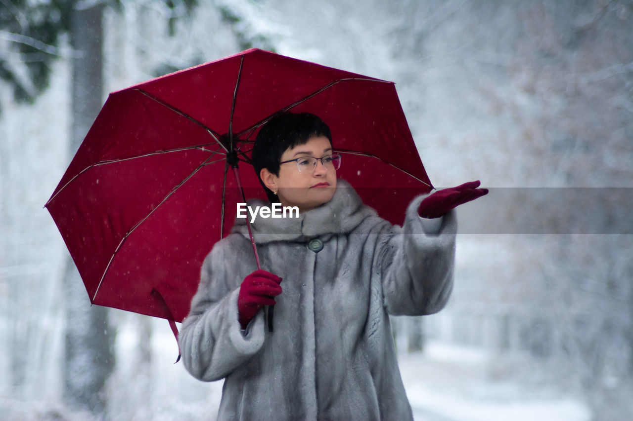 Woman in a gray fur coat with a red umbrella stands on a white, snow-covered background.