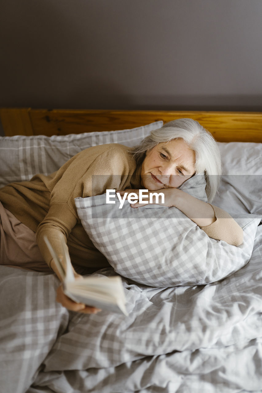Senior woman reading book while lying on bed at home
