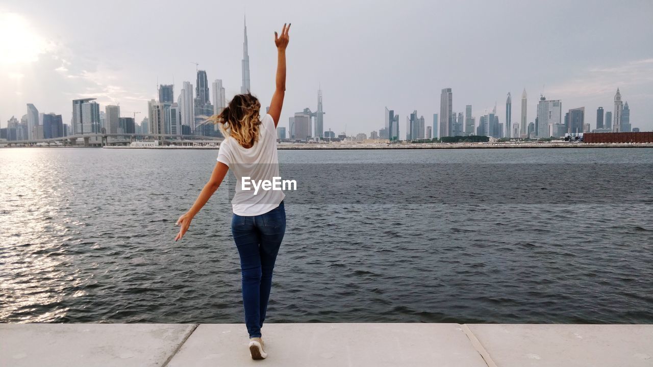Rear view of woman with arm raised standing by sea with urban skyline in background