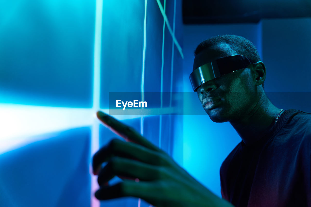 Concentrated young african american male in contemporary vr glasses touching large projector screen with geometric pattern while exploring cyberspace in dark room with neon illumination