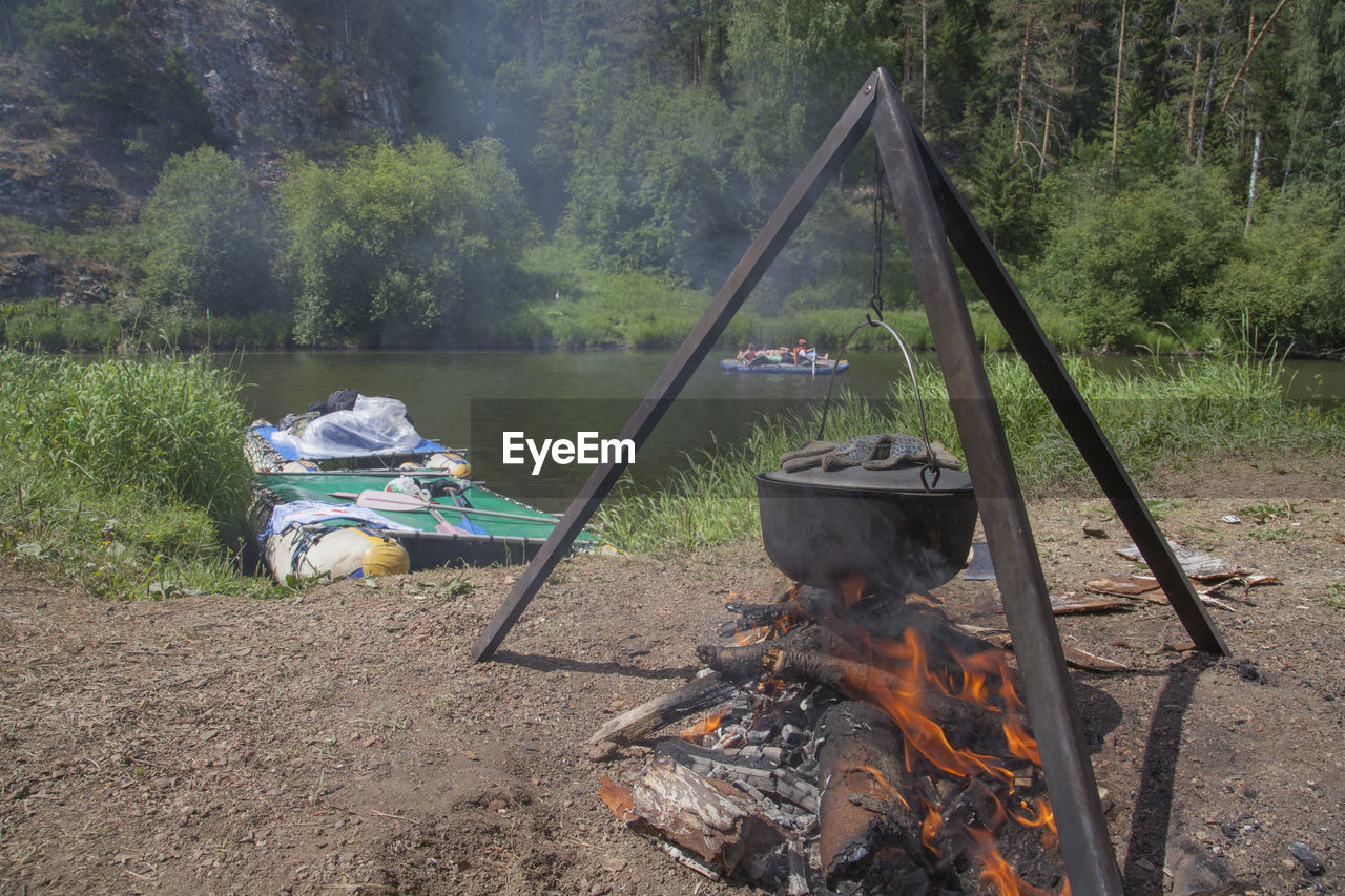 Camping stove hanging over bonfire against river at forest