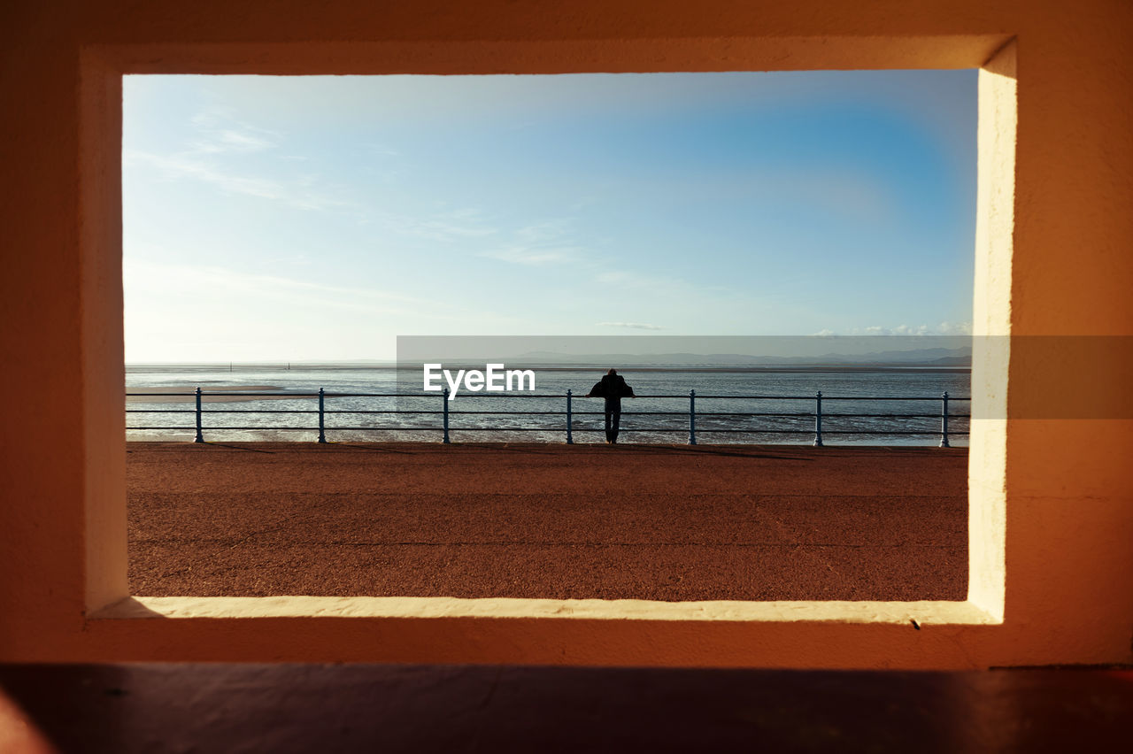 Rear view of man standing on promenade by sea seen through window