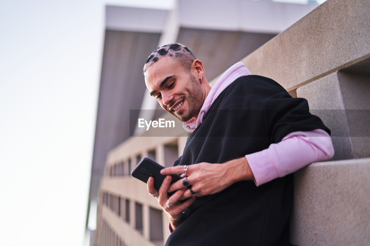 Low angle of trendy young male in hoodie using mobile phone and leaning on concrete wall and smiling widely