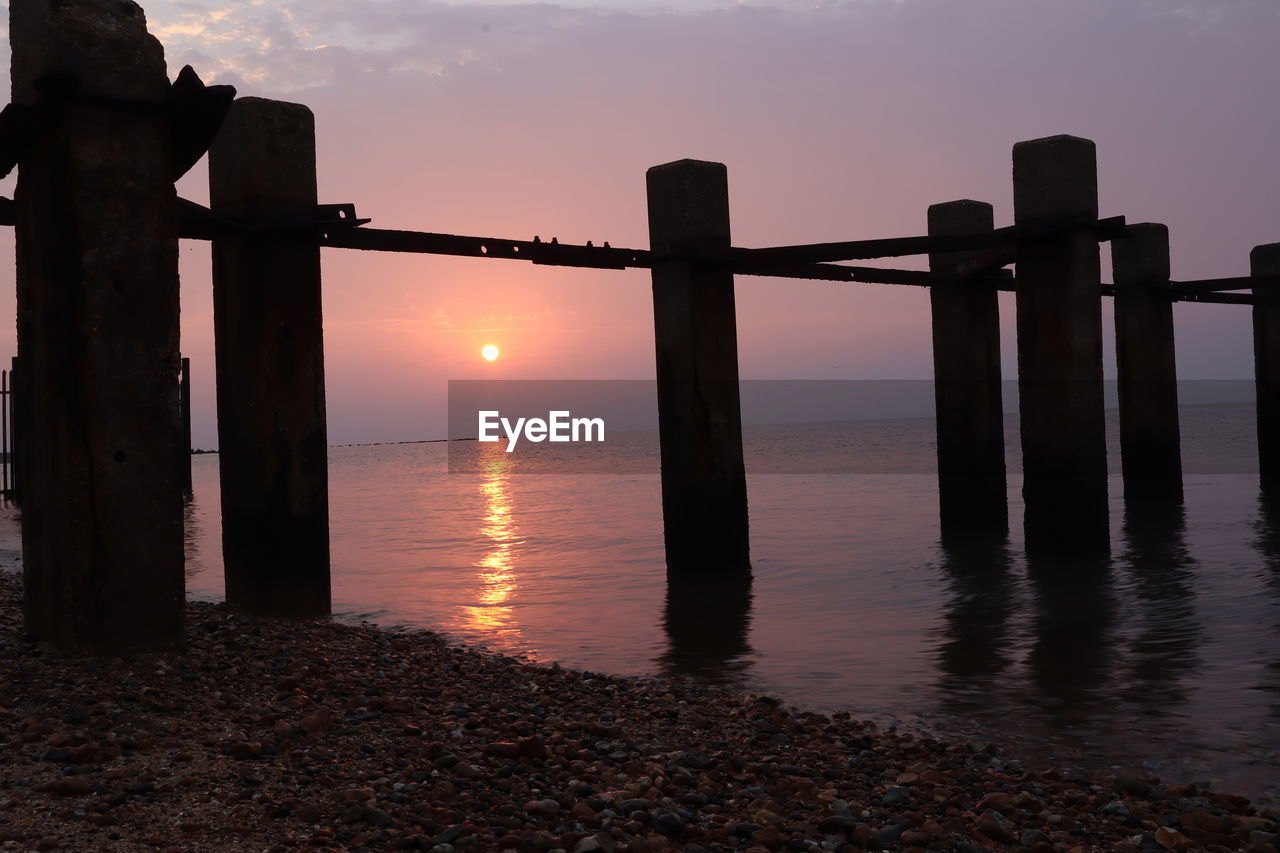 SILHOUETTE WOODEN POSTS ON SEA AGAINST SKY DURING SUNSET