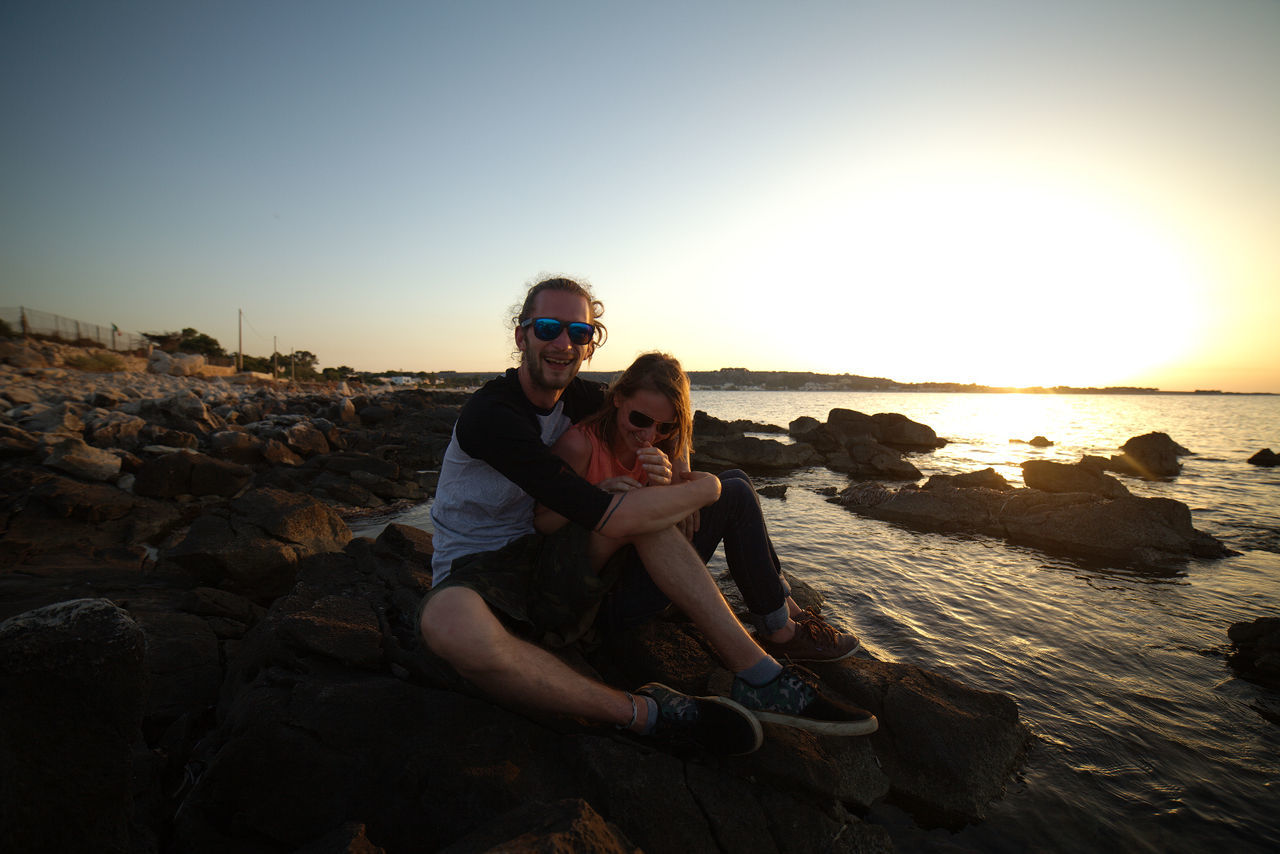 Happy man and woman sitting on rocks at beach against sky during sunset