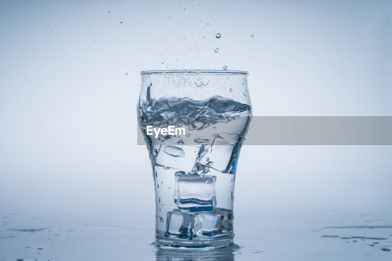 water, refreshment, studio shot, drinking glass, drink, food and drink, glass, household equipment, cold temperature, ice, splashing, nature, no people, drinking water, indoors, freshness, frozen, drop, distilled beverage, freezing, alcoholic beverage, motion, cold drink, transparent, wet, ice cube, colored background, blue, gray background, purity
