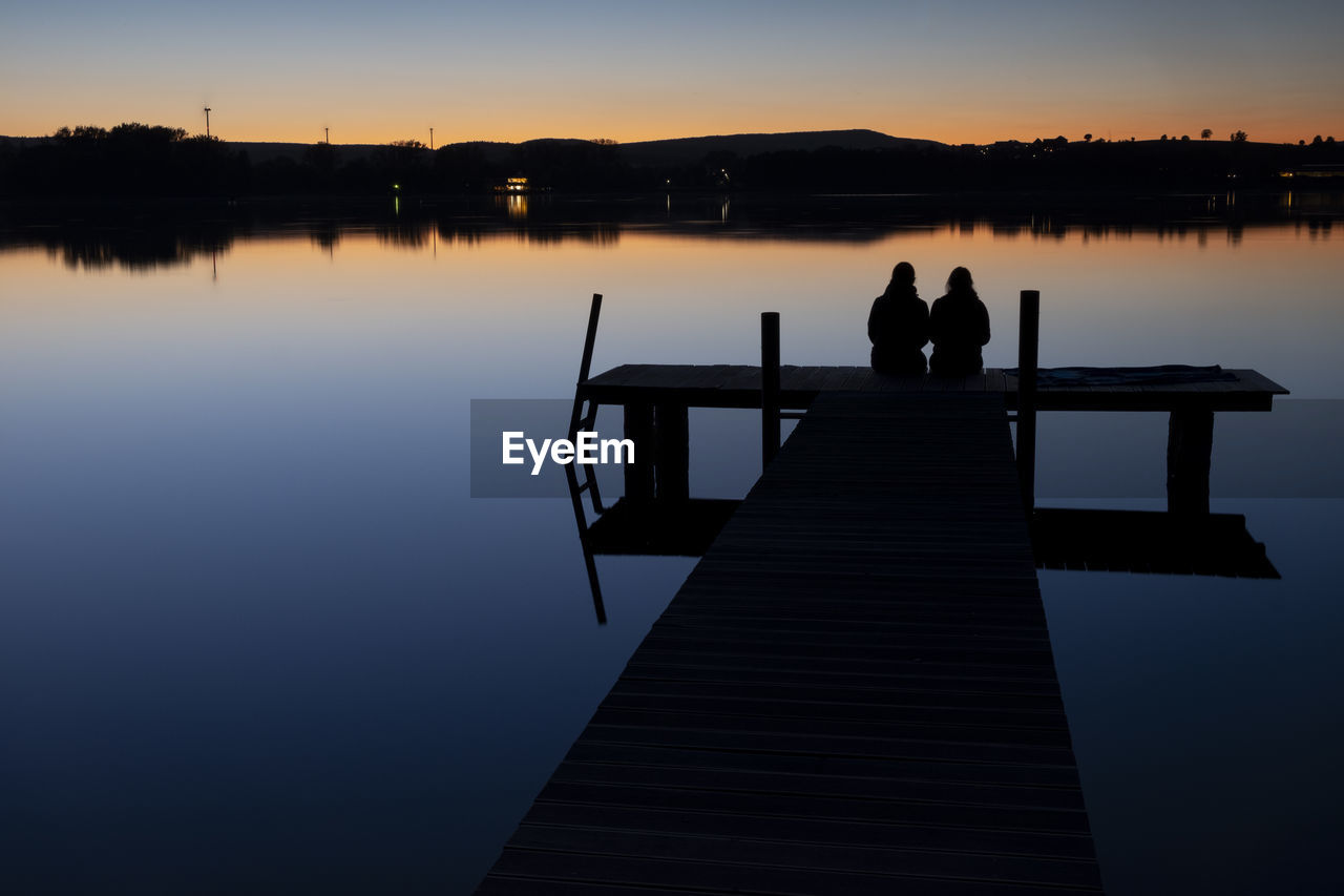 SILHOUETTE PEOPLE SITTING ON PIER AT LAKE AGAINST SKY