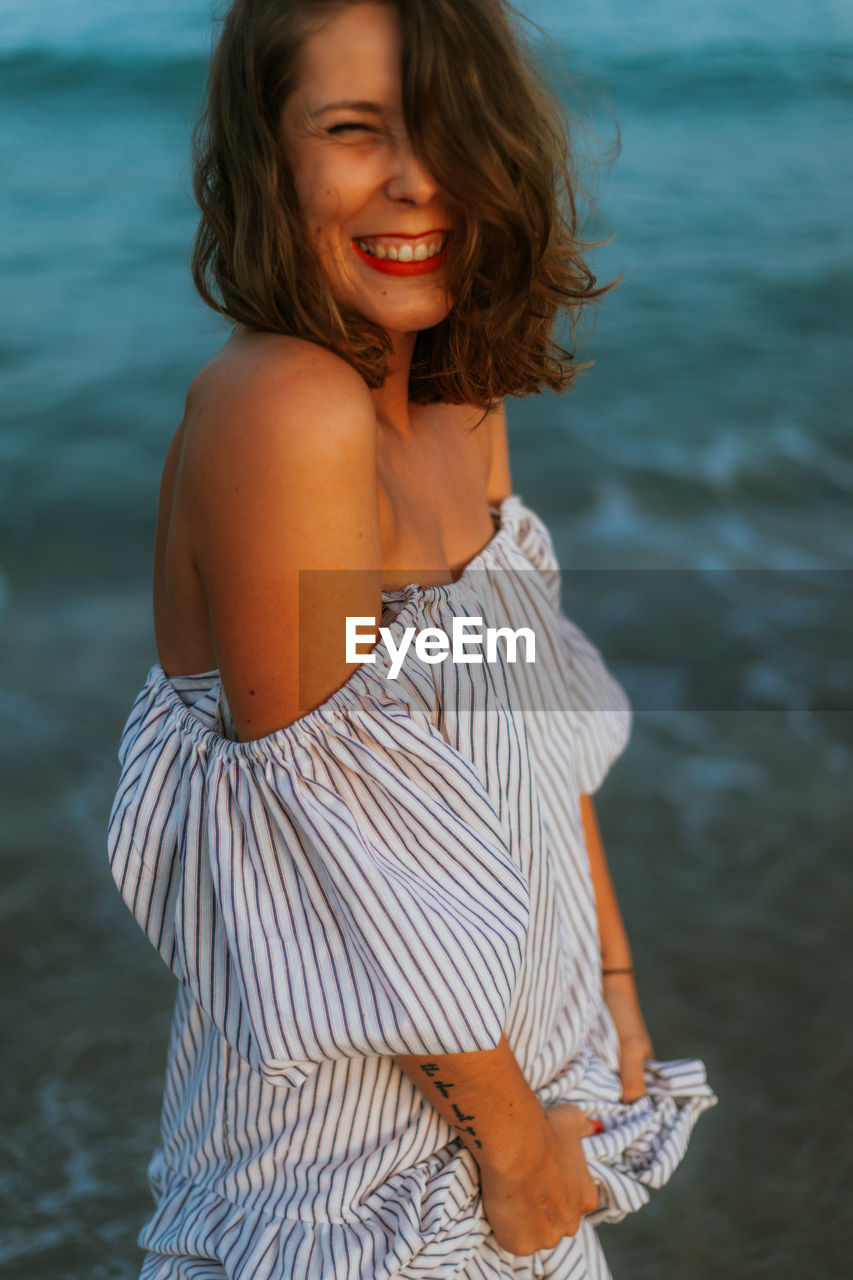 Happy woman in light dress walking among small sea waves on empty coastline at dusk with closed eyes