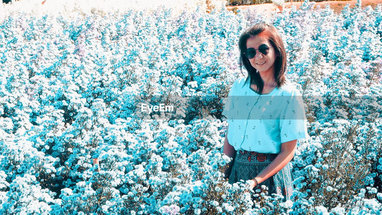 spring, one person, blue, glasses, women, sunglasses, casual clothing, young adult, flower, front view, day, leisure activity, lifestyles, standing, fashion, adult, plant, nature, three quarter length, smiling, portrait, outdoors, happiness, female, looking at camera, emotion, growth, flowering plant, field, person, clothing, hairstyle, sunlight, beauty in nature, land, long hair