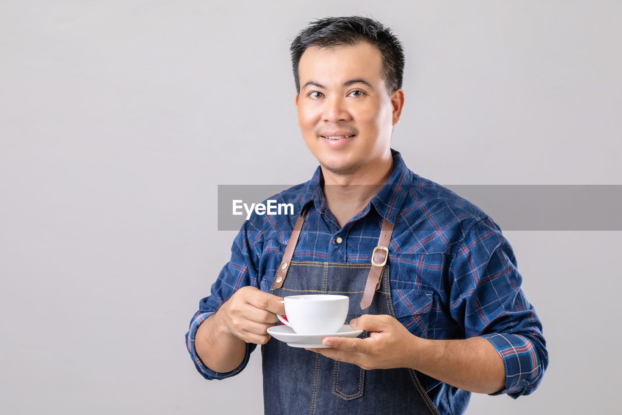 Portrait asian man in barista uniform holding white coffee cup isolated on grey background