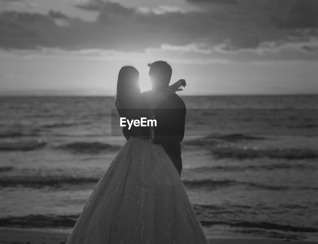 Married couple standing on beach against sky during sunset