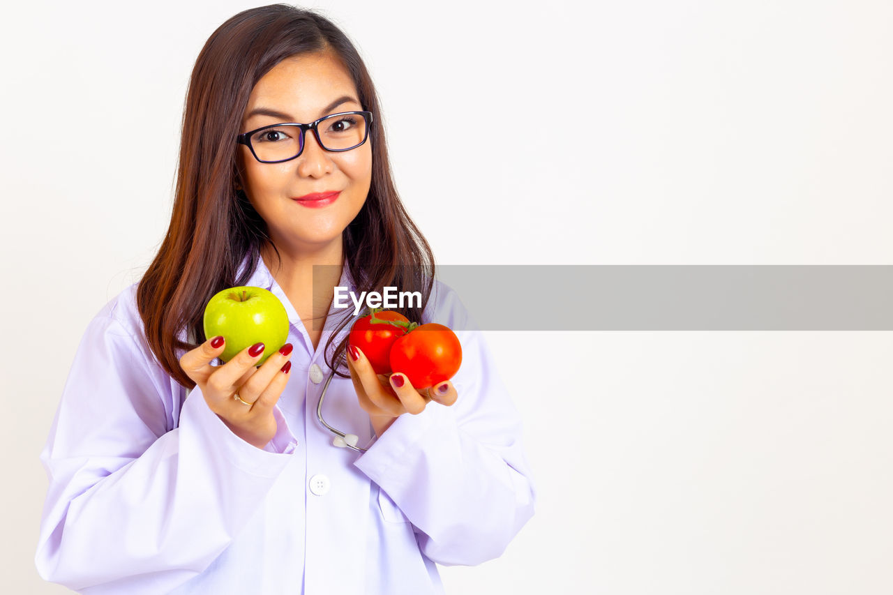 Portrait of smiling doctor holding fruits while standing against white background