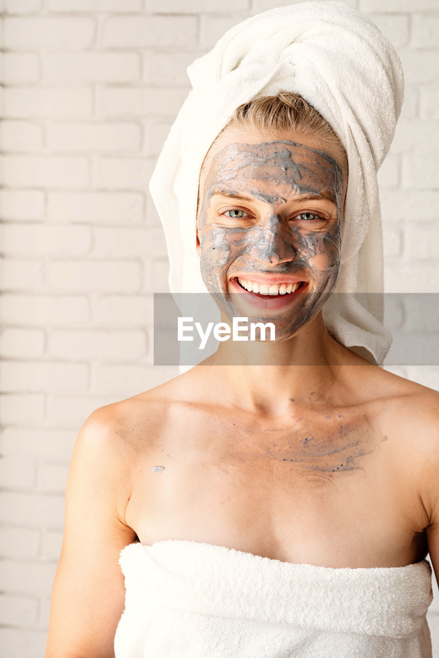 Young smiling woman wearing white bath towels with a clay facial mask on her face