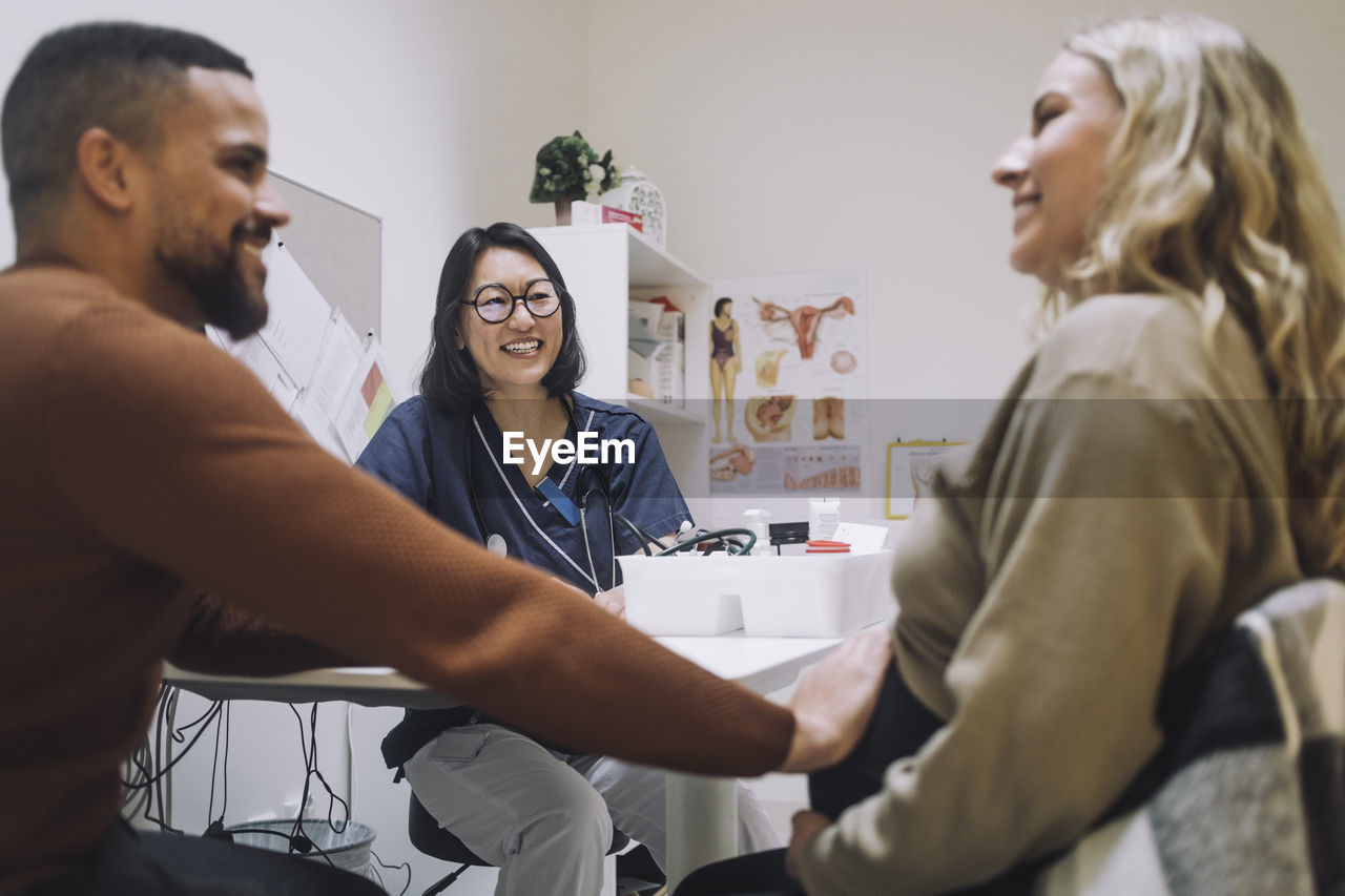 Happy female gynecologist looking at smiling man touching stomach of pregnant woman in clinic