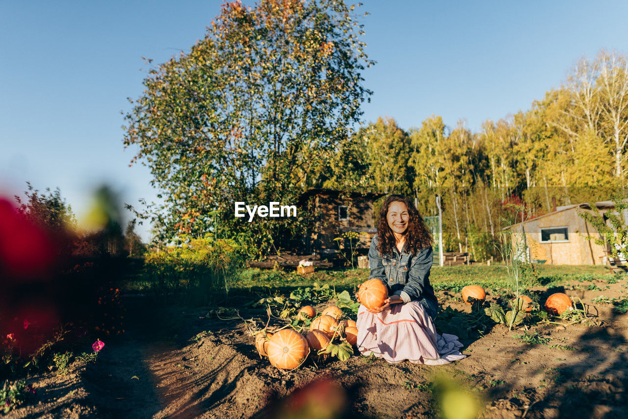Joyful woman in denim jacket, holding pumpkin on sunny day in the countryside