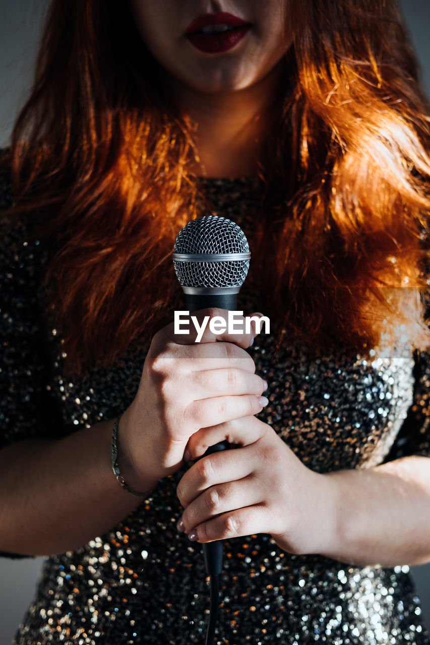 Faceless portrait of redhead woman in sparkly evening dress holding microphone on dark night