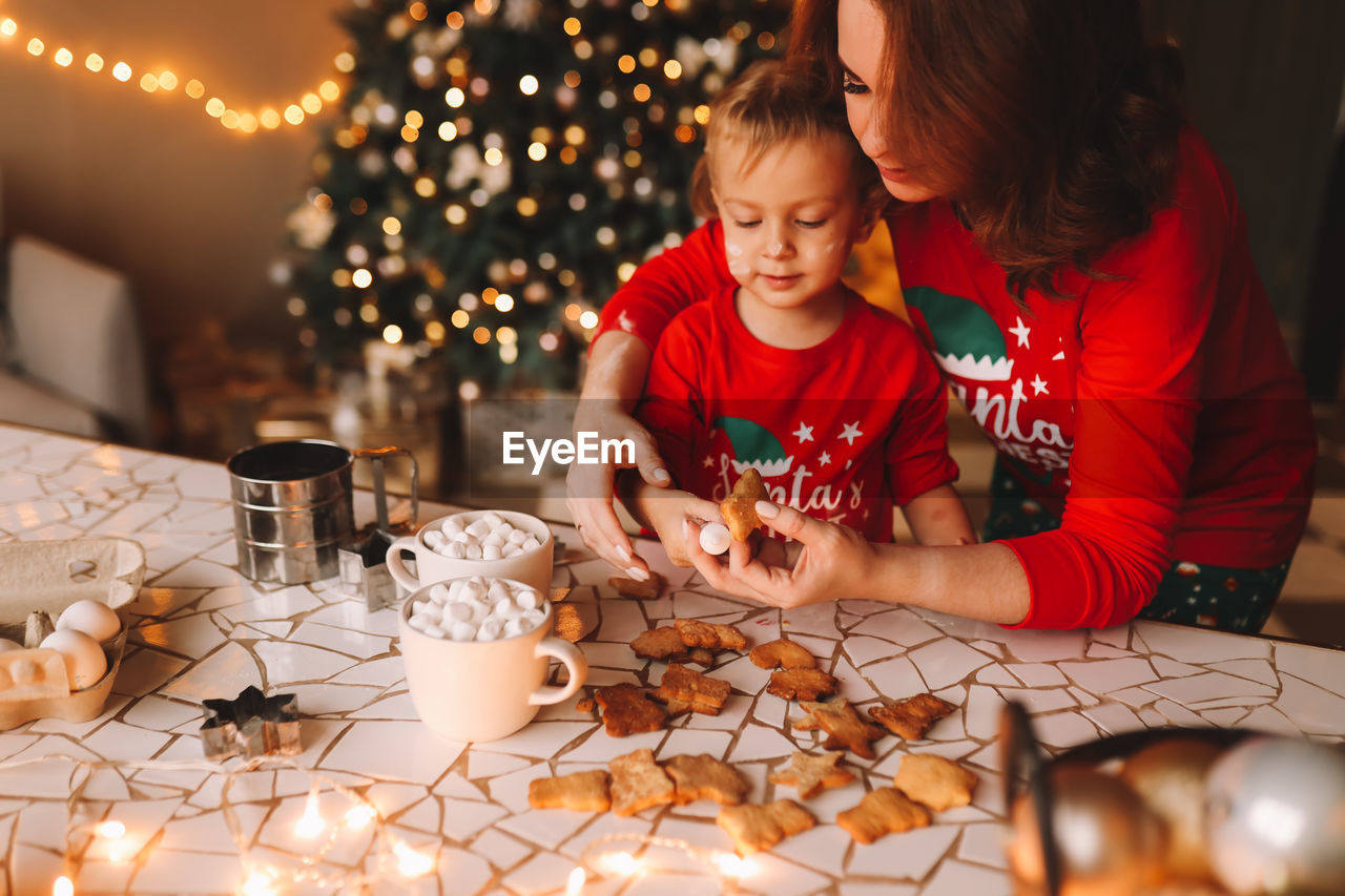 Parents and a child in red pajamas prepare christmas cookies in the decorated kitchen of the house