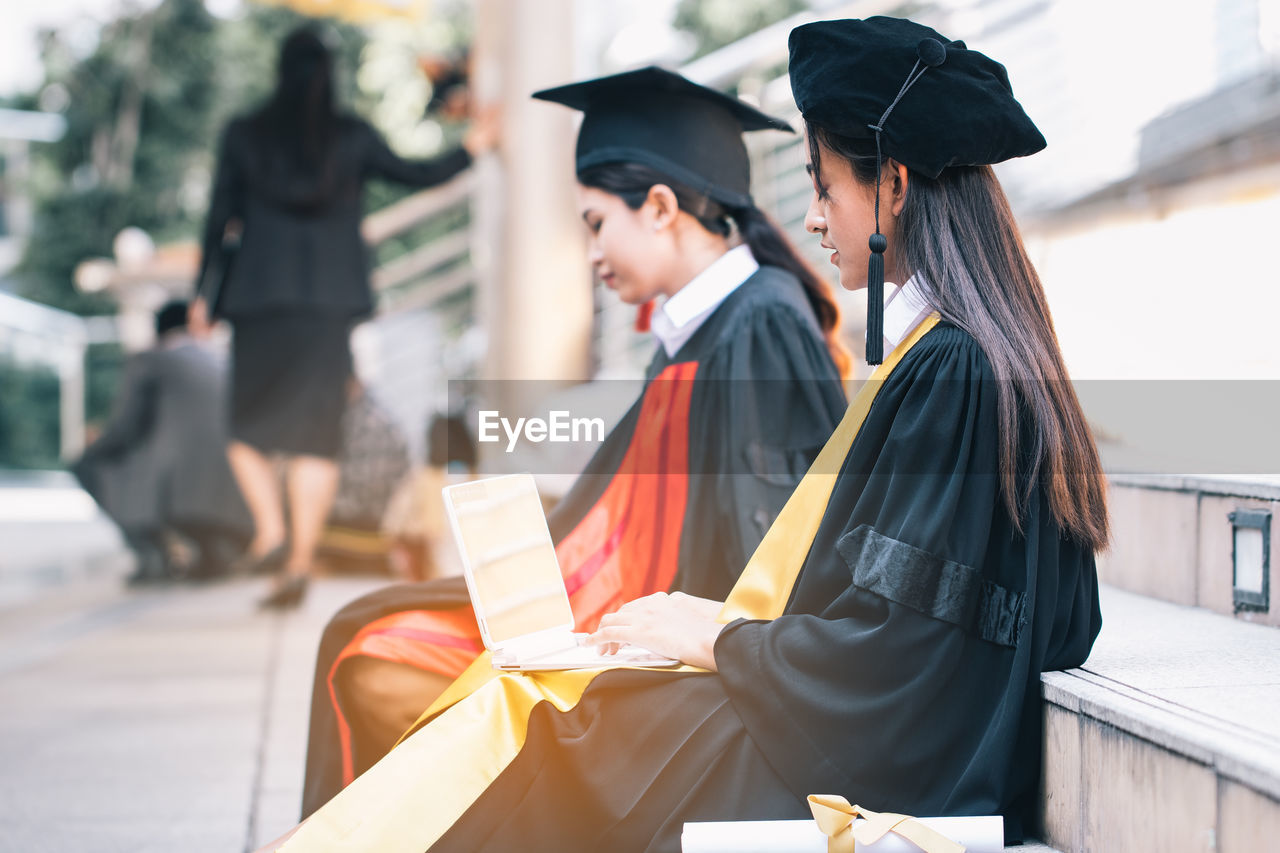 Side view of teenage girl in graduation gown using laptop while sitting on steps