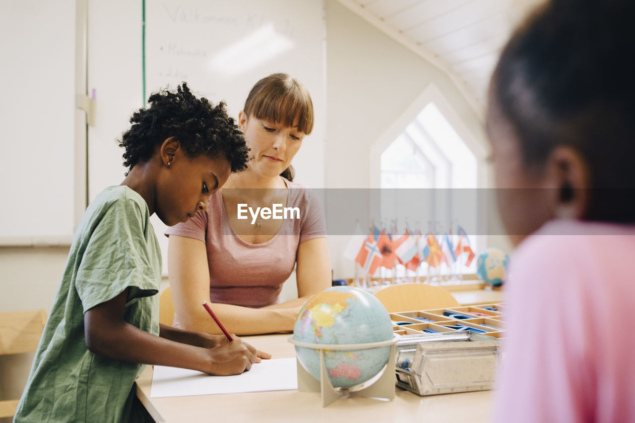 Teacher looking at boy writing on paper at table in classroom