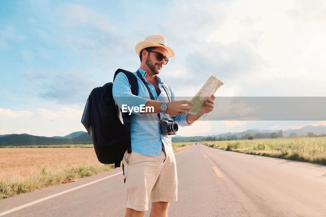 Man wearing hat holding map standing on road against sky