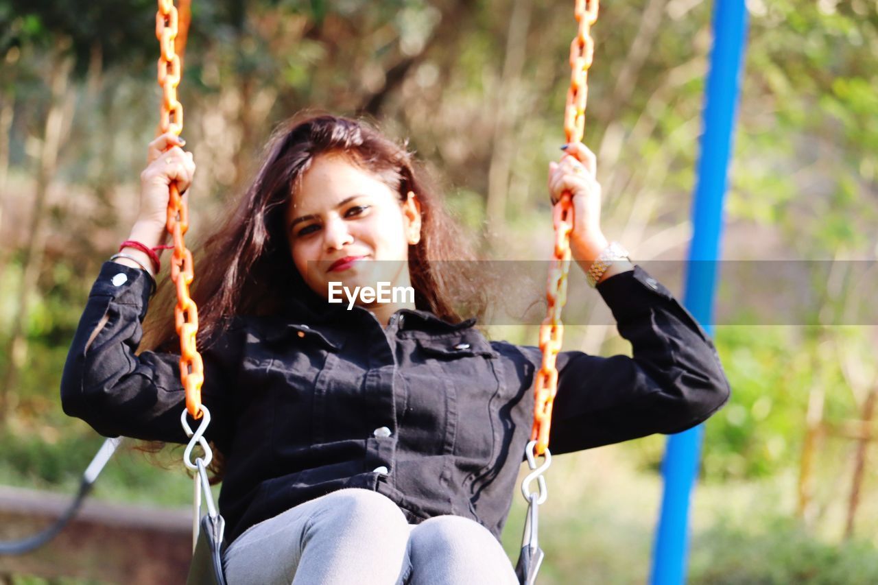 swing, playground, rope, happiness, one person, childhood, smiling, women, leisure activity, nature, child, emotion, chain, fun, hanging, female, safety harness, adventure, enjoyment, portrait, lifestyles, cheerful, joy, sports, positive emotion, looking at camera, sitting, recreation, outdoor play equipment, outdoors, rope swing, park - man made space, tree, adult, relaxation, park, person, spring, day, casual clothing, activity, holding, sunlight, hairstyle, extreme sports, teeth, forest, three quarter length, plant, vitality, young adult, front view, smile, climbing, motion, cute, focus on foreground, carefree, long hair