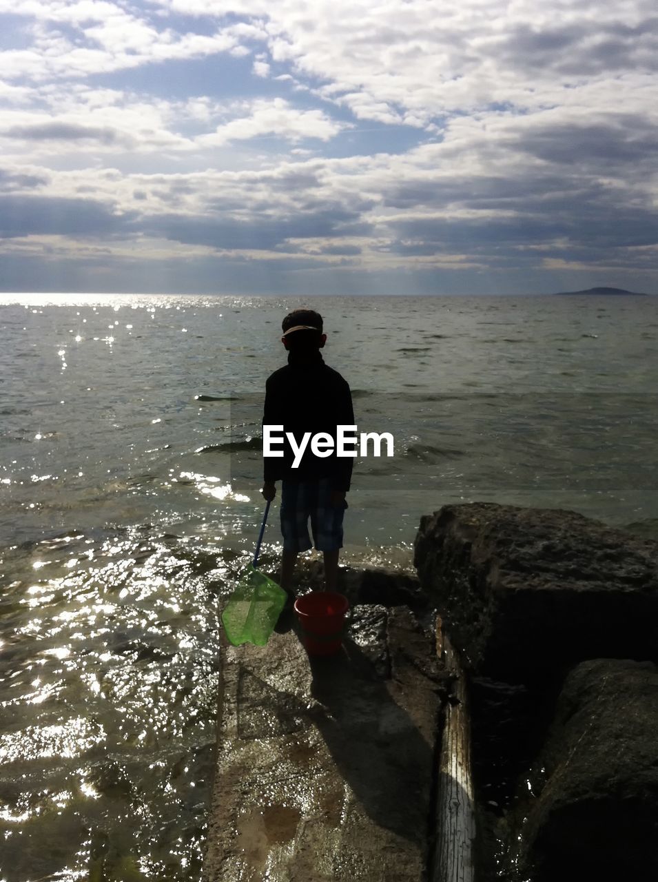 Boy standing on rock by sea against cloudy sky