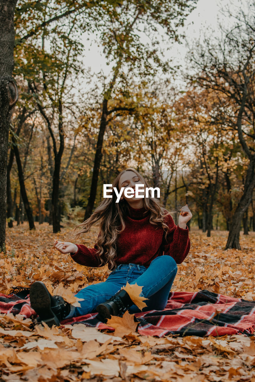 Smiling young woman sitting on autumn leaves against trees in forest