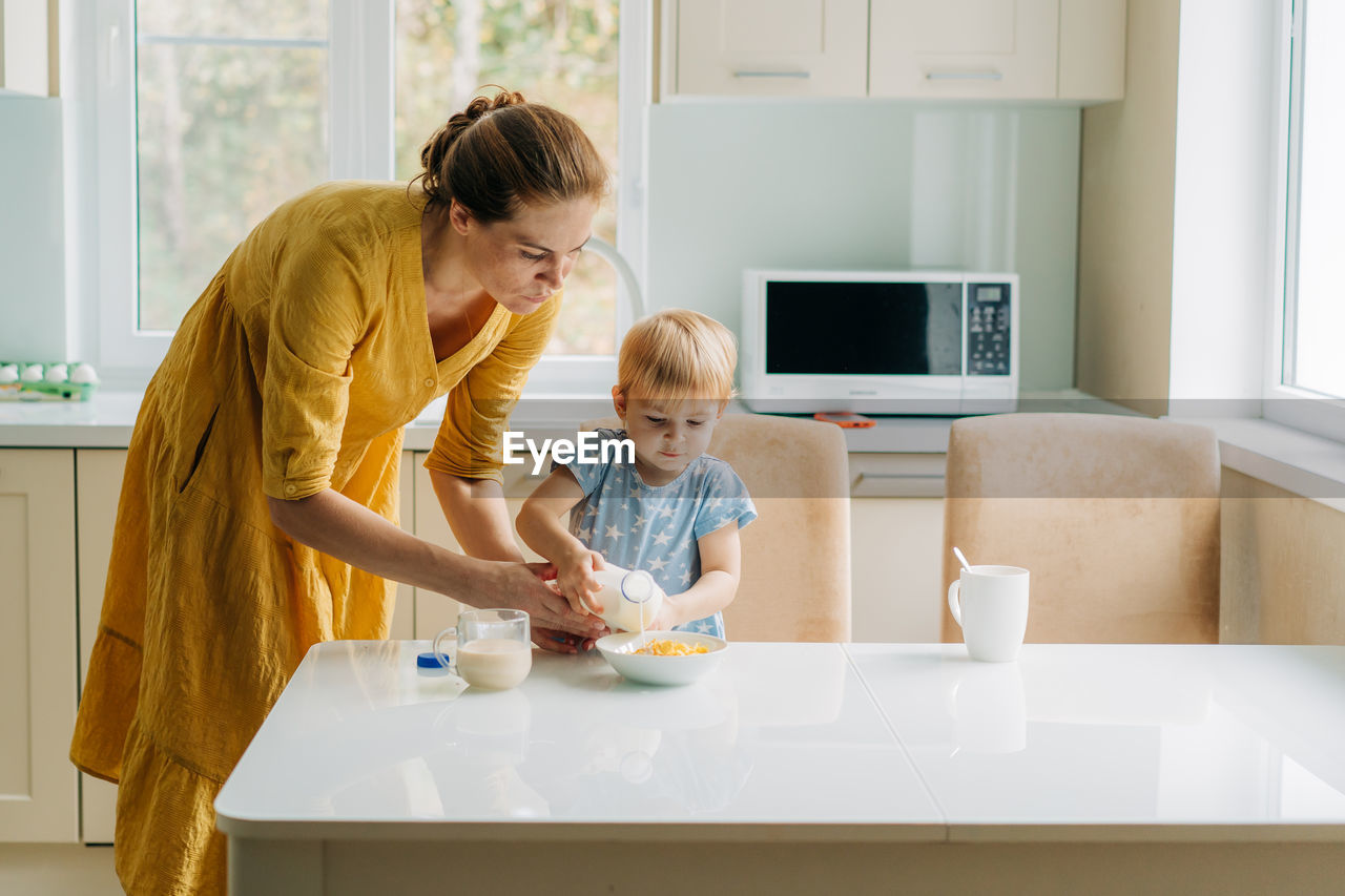 Toddler little daughter and mother cook corn flakes with milk for breakfast.