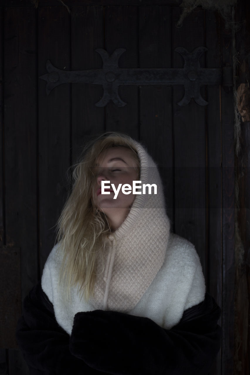 one person, women, clothing, blond hair, darkness, portrait, adult, black, winter, young adult, long hair, emotion, female, entrance, front view, door, waist up, cold temperature, human face, hairstyle, wood, looking at camera, standing, dark, sadness, person, indoors, hood, hood - clothing, child, warm clothing, architecture