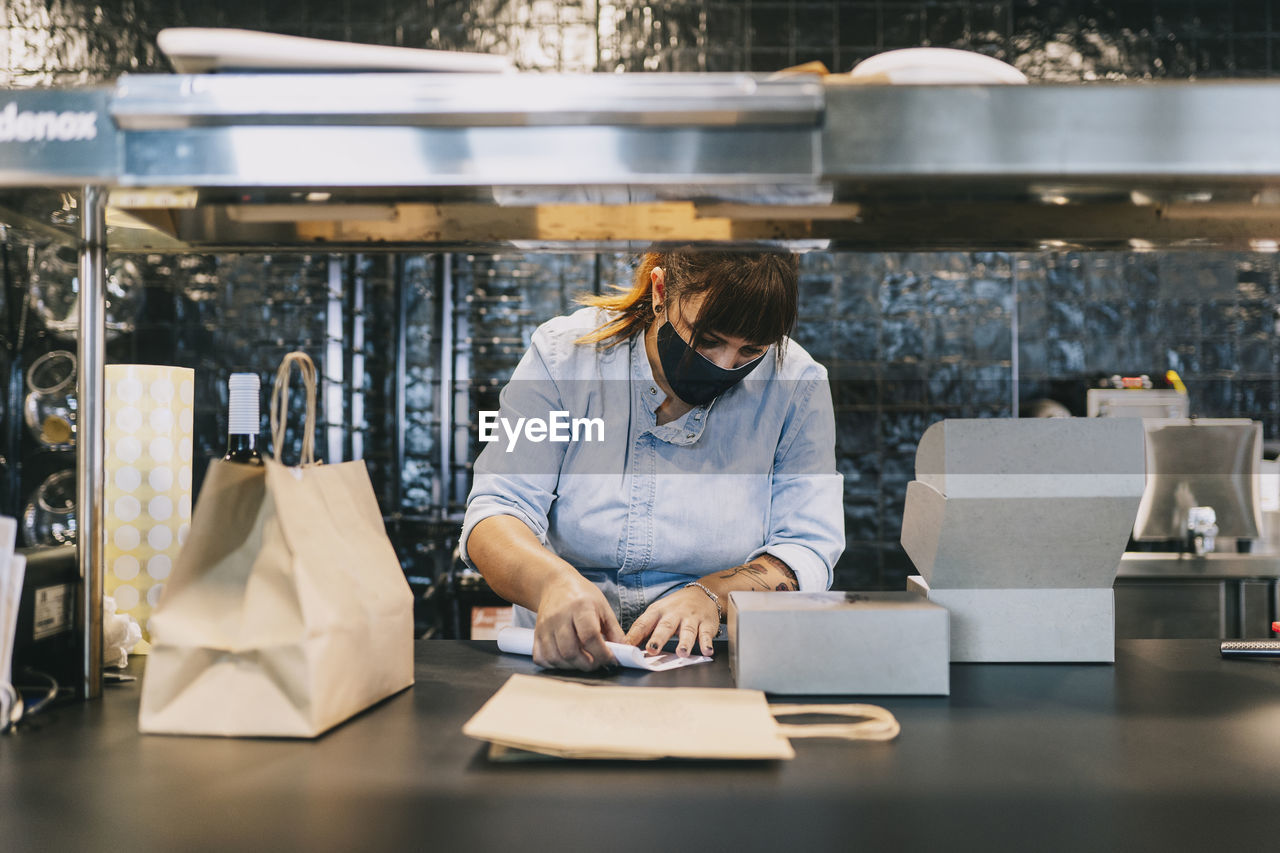 Female chef holding paper by take out containers at restaurant kitchen counter during covid-19