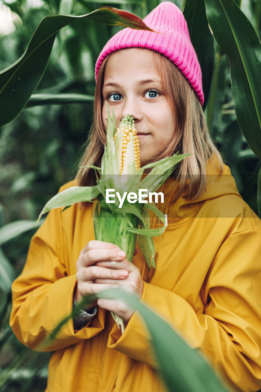 Funny little girl dressed in a yellow raincoat and a hot pink cap spoils and bites corn 