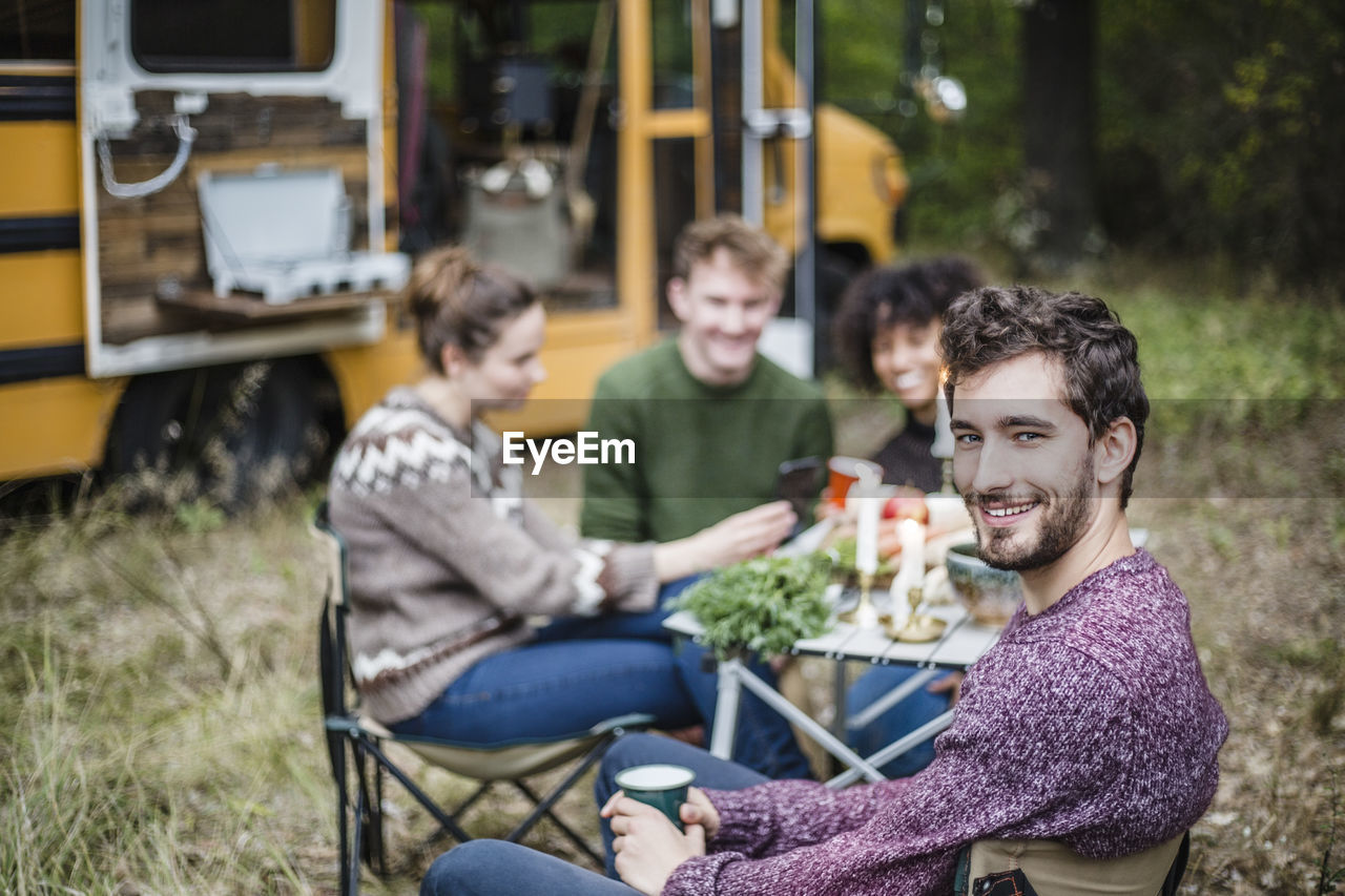 Portrait of smiling man sitting with friends at table during camping in forest