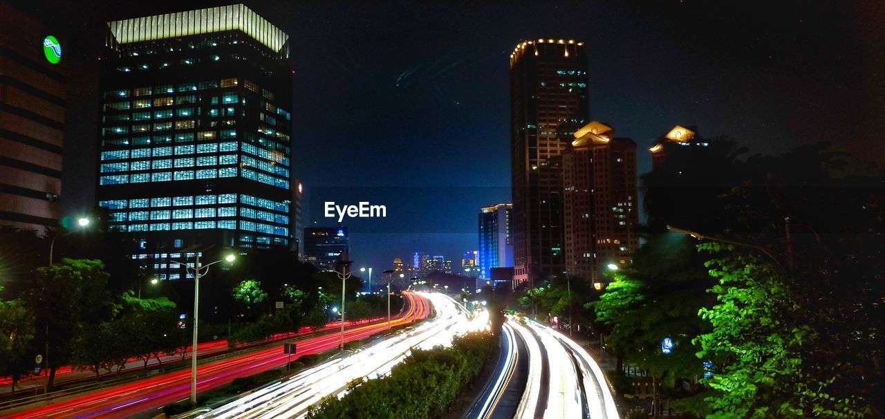 LIGHT TRAILS ON ROAD AMIDST BUILDINGS AGAINST SKY AT NIGHT