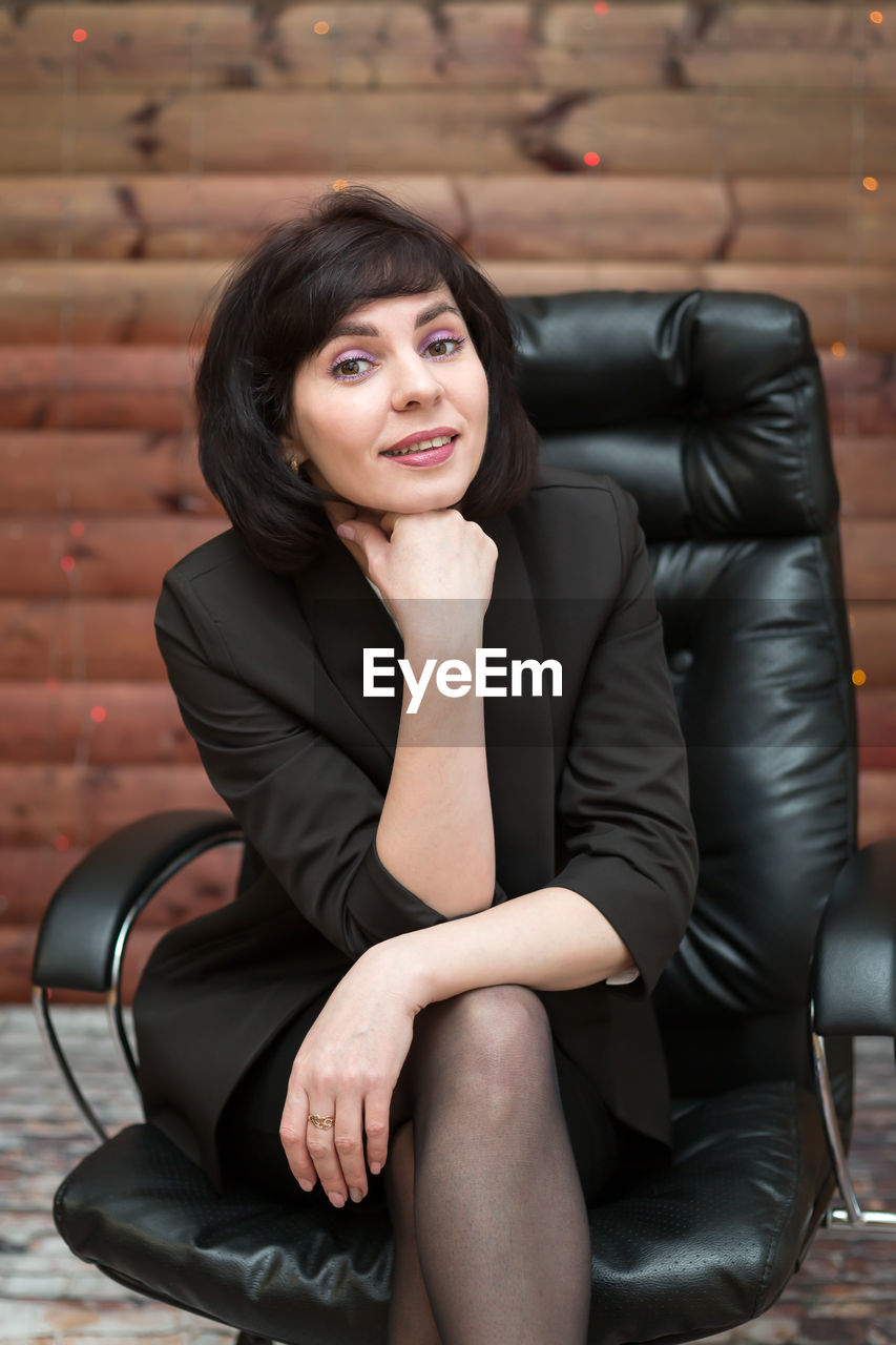 An adult brunette woman in a black formal jacket is sitting in an office chair  wooden background.