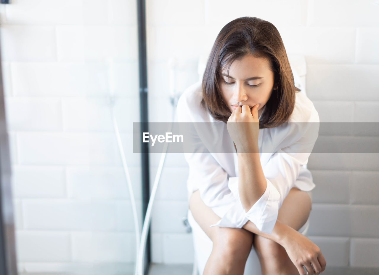 Sad woman sitting on toilet bowl in bathroom at home