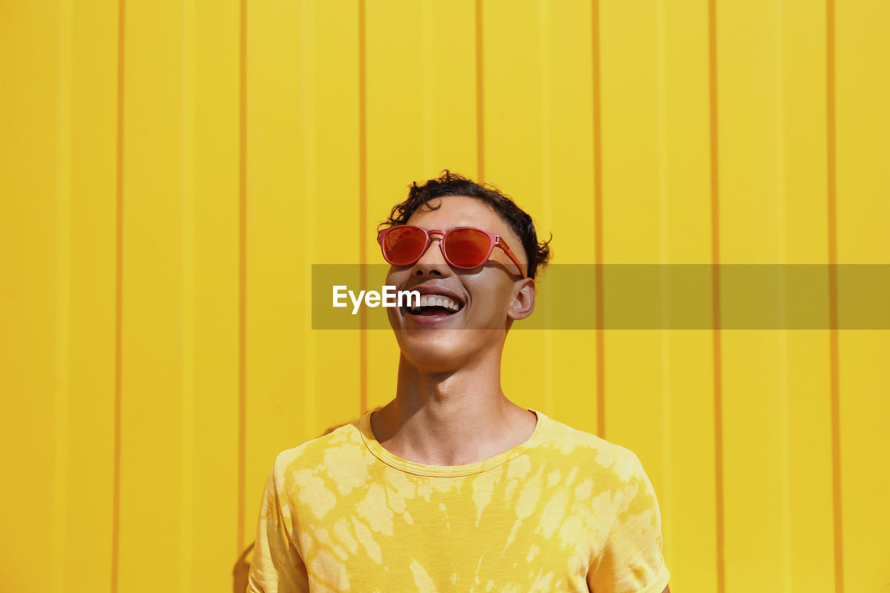 Happy young man wearing red sunglasses in front of yellow wall